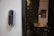 A smart digital lock with a keypad is mounted on a white wall. In the background, there is a blurred view of a dining area with a table and chairs, and some wall art is partially visible.