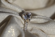 A sparkling diamond ring resting on a textured, shimmering fabric. The ring features a large solitaire diamond set in a prong setting, with smaller diamonds embedded along the band. The fabric underneath is cream-colored with a subtle glitter effect, adding to the overall luxurious ambience.