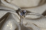 A sparkling diamond ring resting on a textured, shimmering fabric. The ring features a large solitaire diamond set in a prong setting, with smaller diamonds embedded along the band. The fabric underneath is cream-colored with a subtle glitter effect, adding to the overall luxurious ambience.