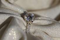 A sparkling diamond ring resting on a textured, shimmering fabric. The ring features a large solitaire diamond set in a prong setting, with smaller diamonds embedded along the band. The fabric underneath is cream-colored with a subtle glitter effect, adding to the overall luxurious ambience.