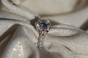 A sparkling diamond ring resting on a textured, shimmering fabric. The ring features a large solitaire diamond set in a prong setting, with smaller diamonds embedded along the band. The fabric underneath is cream-colored with a subtle glitter effect, adding to the overall luxurious ambience.