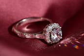 An elegant silver ring is displayed on a deep red silk fabric, featuring a large central clear gemstone surrounded by smaller sparkling stones. The light reflects off the ring creating colorful reflections on the fabric.