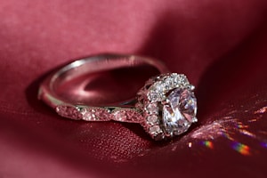 An elegant silver ring is displayed on a deep red silk fabric, featuring a large central clear gemstone surrounded by smaller sparkling stones. The light reflects off the ring creating colorful reflections on the fabric.