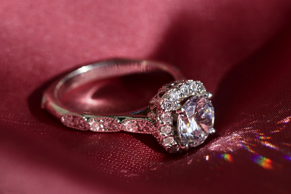 An elegant silver ring is displayed on a deep red silk fabric, featuring a large central clear gemstone surrounded by smaller sparkling stones. The light reflects off the ring creating colorful reflections on the fabric.