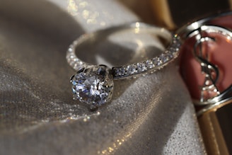 A sparkling diamond ring displayed on a fabric background with a soft, shimmering texture. The ring has a large central gemstone surrounded by smaller diamonds set along the band. The fabric underneath catches the light, adding a luxurious and elegant atmosphere.