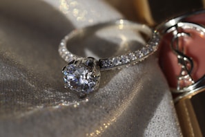 A sparkling diamond ring displayed on a fabric background with a soft, shimmering texture. The ring has a large central gemstone surrounded by smaller diamonds set along the band. The fabric underneath catches the light, adding a luxurious and elegant atmosphere.