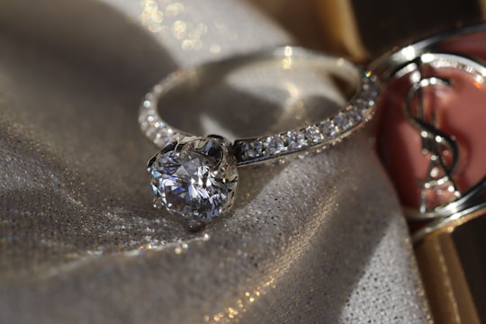 A sparkling diamond ring displayed on a fabric background with a soft, shimmering texture. The ring has a large central gemstone surrounded by smaller diamonds set along the band. The fabric underneath catches the light, adding a luxurious and elegant atmosphere.