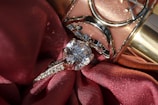 A close-up of a diamond ring resting on rich, red fabric with a shiny metallic object in the background. The ring features a prominent, sparkling diamond set in a silver band with smaller stones embedded along the sides.