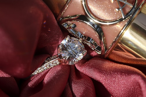 A close-up of a diamond ring resting on rich, red fabric with a shiny metallic object in the background. The ring features a prominent, sparkling diamond set in a silver band with smaller stones embedded along the sides.