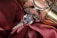 A close-up of a diamond ring resting on rich, red fabric with a shiny metallic object in the background. The ring features a prominent, sparkling diamond set in a silver band with smaller stones embedded along the sides.