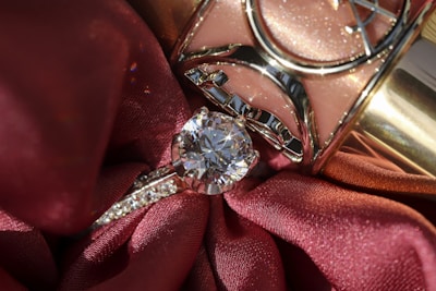 A close-up of a diamond ring resting on rich, red fabric with a shiny metallic object in the background. The ring features a prominent, sparkling diamond set in a silver band with smaller stones embedded along the sides.