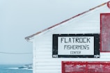 A wooden building with visible white siding and a sign reading 'Flatrock Fishermen's Center'. The structure features red trimming and details, situated near a cold, foggy seascape.