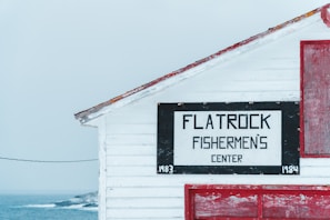 A wooden building with visible white siding and a sign reading 'Flatrock Fishermen's Center'. The structure features red trimming and details, situated near a cold, foggy seascape.