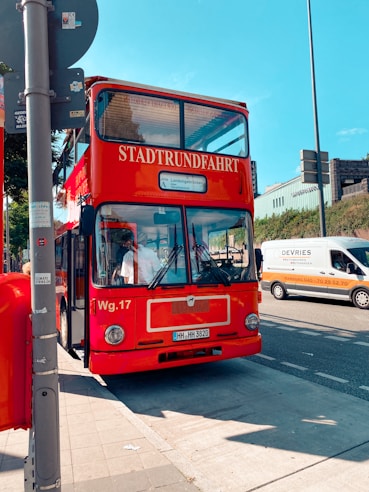 A red double-decker tour bus labeled 'Stadtrundfahrt' is parked by the side of a street under a clear blue sky. The bus is positioned on a city sidewalk, with nearby road traffic including a white and orange van. Various street signs and a pole are visible in the foreground.