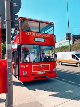 A red double-decker tour bus labeled 'Stadtrundfahrt' is parked by the side of a street under a clear blue sky. The bus is positioned on a city sidewalk, with nearby road traffic including a white and orange van. Various street signs and a pole are visible in the foreground.
