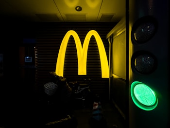 A large, illuminated yellow letter 'M' is prominently displayed against a dark background suggesting the recognizable McDonald's logo. To the right, a green traffic signal light is visible. Below the letter 'M', there are various objects including a basket and parts of a bicycle.