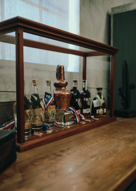 A display case on a wooden surface contains various bottles of spirits, each adorned with medals. The centerpiece is a large, ornate trophy and several award ribbons. The background features a softly lit window and a textured wall, contributing to a vintage and sophisticated atmosphere.