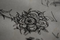 A detailed pen or pencil sketch of a floral design, focusing on intricately drawn roses with layered petals. The lines are bold and abstract, creating a textured appearance against a plain, off-white background.