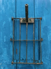 A weathered metal bracket with a vertical hollow pipe is mounted against a distressed blue wooden surface. The bracket appears rusty and old, standing out against the vivid blue backdrop with peeling paint.