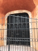 A large, heavy black metal gate is set into a stone archway surrounded by textured, warm orange walls. Intricate decorative metalwork can be observed on the gate, with a pattern of circles and vertical bars. The stone archway has an aged, weathered appearance.