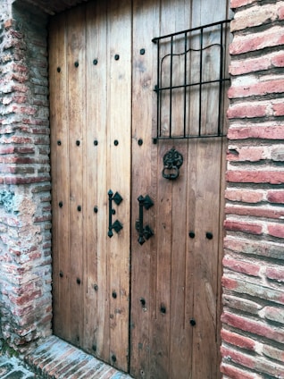 A wooden door with metal studs set into a brick wall, featuring two ornate black metal handles and a decorative grille across a small window.