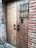 A wooden door with metal studs set into a brick wall, featuring two ornate black metal handles and a decorative grille across a small window.