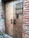 A wooden door with metal studs set into a brick wall, featuring two ornate black metal handles and a decorative grille across a small window.
