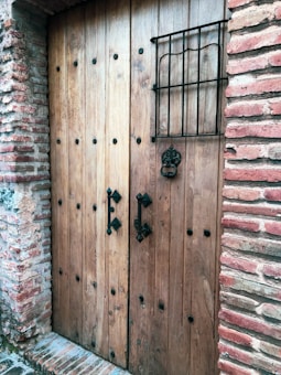 A wooden door with metal studs set into a brick wall, featuring two ornate black metal handles and a decorative grille across a small window.