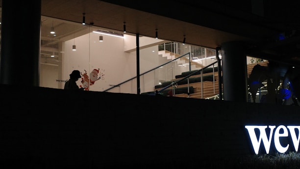 The image depicts a dimly lit interior space viewed from the outside. Large glass windows reveal an interior decorated with hanging lights and a staircase with wooden steps. The silhouette of a person can be seen against the backdrop of the building's entrance, which features the prominent illuminated signage 'wework'. The ambiance inside is warm with modern furnishings.