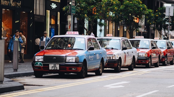 Several taxis in a row are parked along a busy urban street lined with modern buildings and shops. Pedestrians are walking on the sidewalk, and trees can be seen lining the street.
