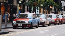 Several taxis in a row are parked along a busy urban street lined with modern buildings and shops. Pedestrians are walking on the sidewalk, and trees can be seen lining the street.