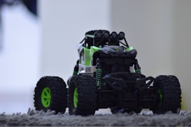 A toy or model off-road vehicle with large, rugged tires and a bold green and black design sits on a textured carpeted surface. The vehicle features intricate details such as shock absorbers and front lights, giving it a realistic appearance. The background is blurred, focusing attention on the vehicle.