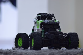A toy or model off-road vehicle with large, rugged tires and a bold green and black design sits on a textured carpeted surface. The vehicle features intricate details such as shock absorbers and front lights, giving it a realistic appearance. The background is blurred, focusing attention on the vehicle.