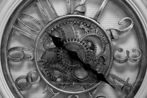 An intricate clock face with Roman numerals and visible gear mechanisms inside. The numbers and gears are metallic with a rustic, antique finish. Two ornate clock hands point towards different numbers, adding to the vintage aesthetic.