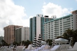 A row of high-rise buildings with modern architecture rises against a backdrop of a partly cloudy sky. In the foreground, striped umbrellas and white lounge chairs are positioned, suggesting a beach or poolside setting. Palm trees are visible around the area, enhancing the resort-like atmosphere.
