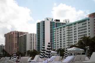 A row of high-rise buildings with modern architecture rises against a backdrop of a partly cloudy sky. In the foreground, striped umbrellas and white lounge chairs are positioned, suggesting a beach or poolside setting. Palm trees are visible around the area, enhancing the resort-like atmosphere.