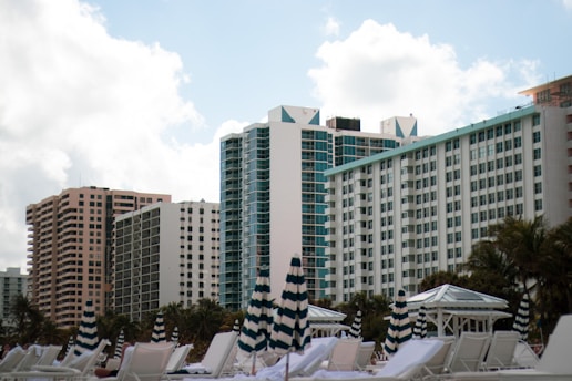 A row of high-rise buildings with modern architecture rises against a backdrop of a partly cloudy sky. In the foreground, striped umbrellas and white lounge chairs are positioned, suggesting a beach or poolside setting. Palm trees are visible around the area, enhancing the resort-like atmosphere.