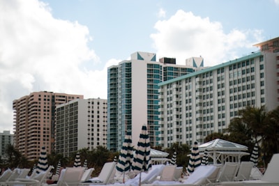 A row of high-rise buildings with modern architecture rises against a backdrop of a partly cloudy sky. In the foreground, striped umbrellas and white lounge chairs are positioned, suggesting a beach or poolside setting. Palm trees are visible around the area, enhancing the resort-like atmosphere.