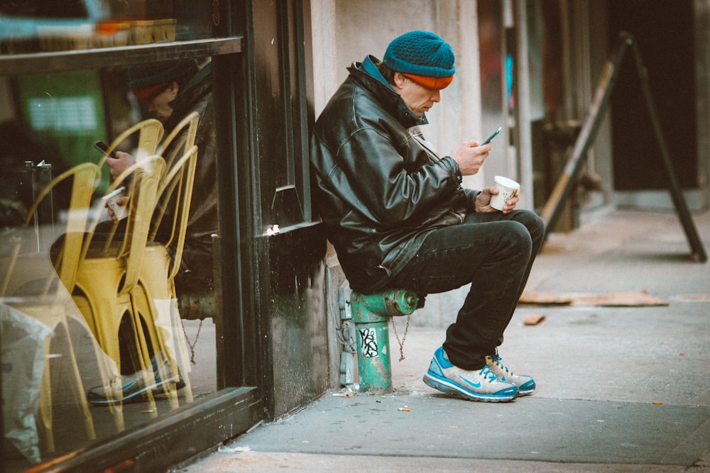 Hombre con chaqueta de cuero negro y gorro de punto azul sentado en el suelo de hormigón gris