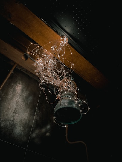 A hanging industrial-style light fixture adorned with a cascade of fairy lights. The lights create a warm, enchanting glow against a dark background, highlighting the rustic texture of the metal fixture and the wooden beam it is attached to.