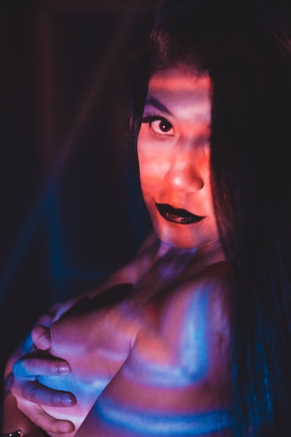 A person with long black hair is illuminated by vibrant lighting that casts red and blue shadows across their face and body. They have dark eye makeup and bold black lipstick, gazing directly at the camera with a captivating expression. The lighting creates a dramatic and mysterious atmosphere.