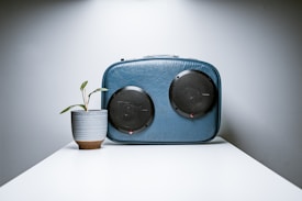 A minimalist scene featuring a stylish blue suitcase with two round black speakers, positioned on a white table. Next to the suitcase is a small green plant in a ceramic pot. The lighting is soft, casting gentle shadows, and the overall aesthetic is modern and clean.