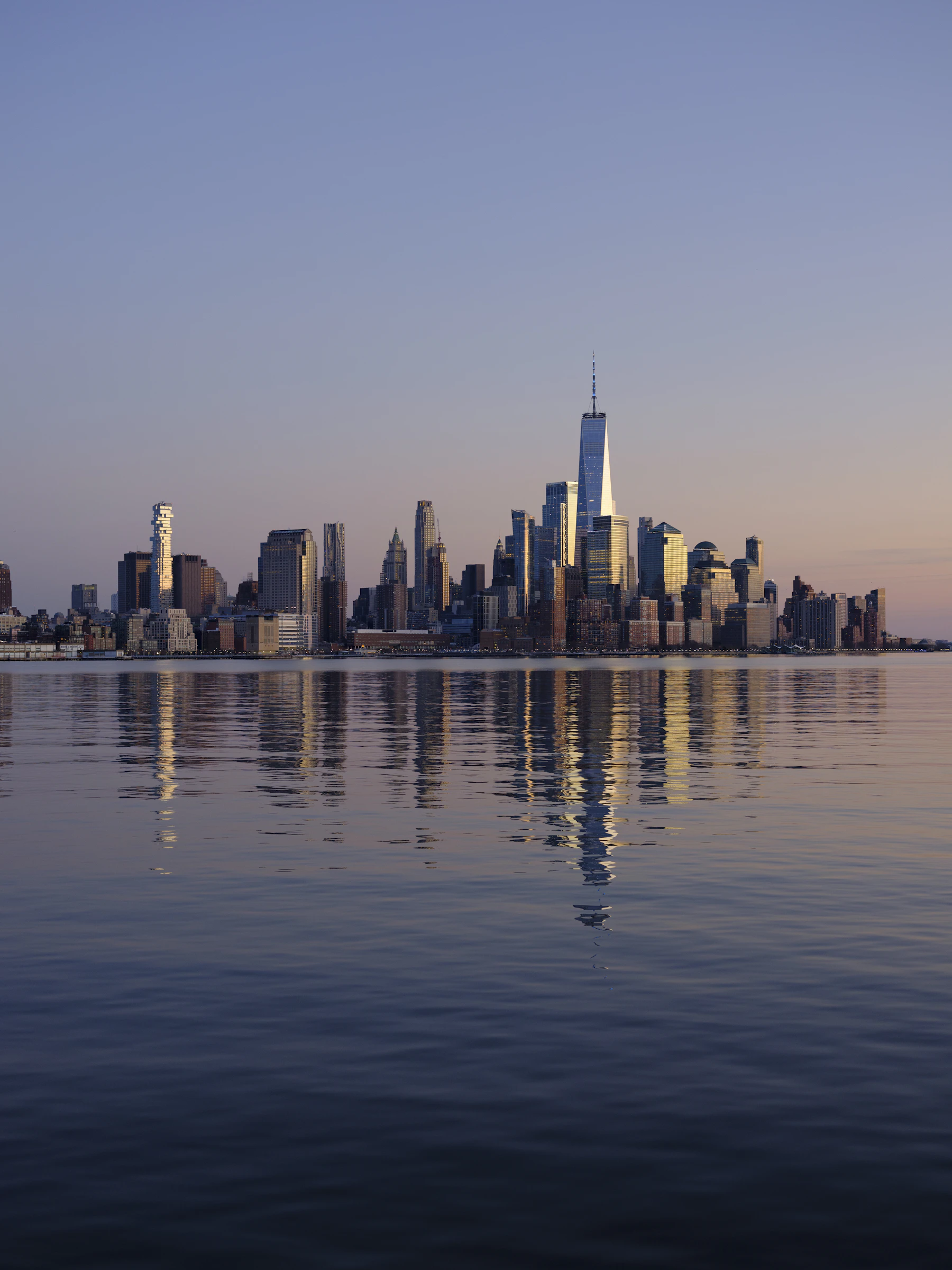 Travel photograph: Manhattan’s southern skyline seen across practical Hudson River water in soft evening light.