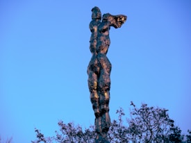A tall, abstract sculpture resembling a human form extends upwards against a clear blue sky. The reflective surface of the sculpture contrasts with the vibrant sky, and the sculpture itself appears to be made of a dark, textured material. To the lower right, part of the frame is filled by the branches of trees, adding an earthy contrast to the scene.