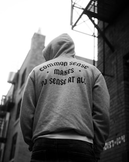 A person wearing a hooded sweatshirt with the phrase 'Common sense makes no sense at all' is standing in an urban setting. The focus is on the back of the sweatshirt, with surrounding buildings slightly out of focus.
