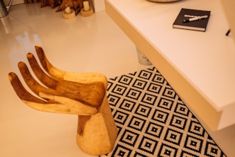 A wooden chair shaped like a hand next to a desk. The floor is covered with a black and white geometric patterned rug. On the white desk, there is a notebook with a pen placed on top. Some decorative items and candles are seen in the background.