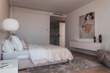 A modern bedroom features a neatly made bed with white and beige bedding, flanked by pillows. A large, round lamp hangs above a bedside table that holds a few items. An abstract painting decorates the wall above a sleek white console, which holds two black vases. The room has a soft, plush rug on wooden flooring and an open door leading to another area.