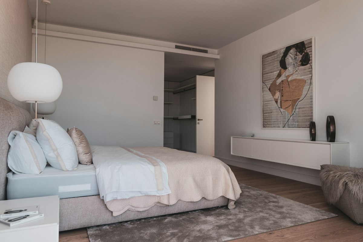 Dormitorio luminoso con paredes blancas, cama con sábanas blancas y manta clara, lámpara de globo colgante, cuadro grande en la pared y piso de madera con alfombra gris