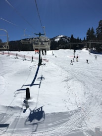 A snowy ski resort with a chairlift in the foreground stretching toward the mountain in the distance. There are several skiers on the slopes, and the scene includes pine trees and mountain peaks under a clear blue sky.