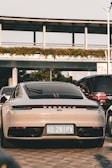 A luxury sports car is parked on a paved surface, positioned in front of a modern structure with greenery along the top. It is surrounded by other vehicles, indicating an urban setting. The car is sleek and shiny, with a focus on its rear design and a visible license plate.